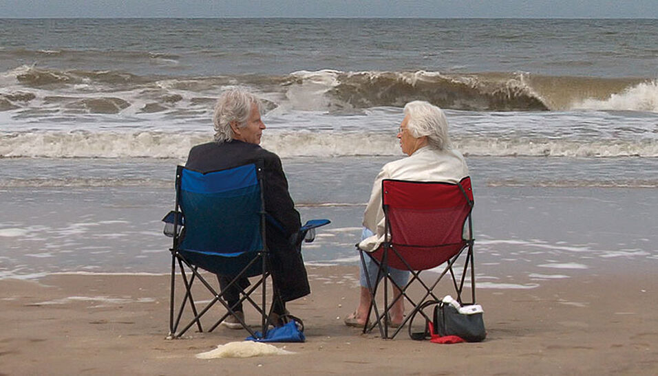 Ein älterer Mann und eine ältere Frau sitzen auf Campingstühlen am Strand.
