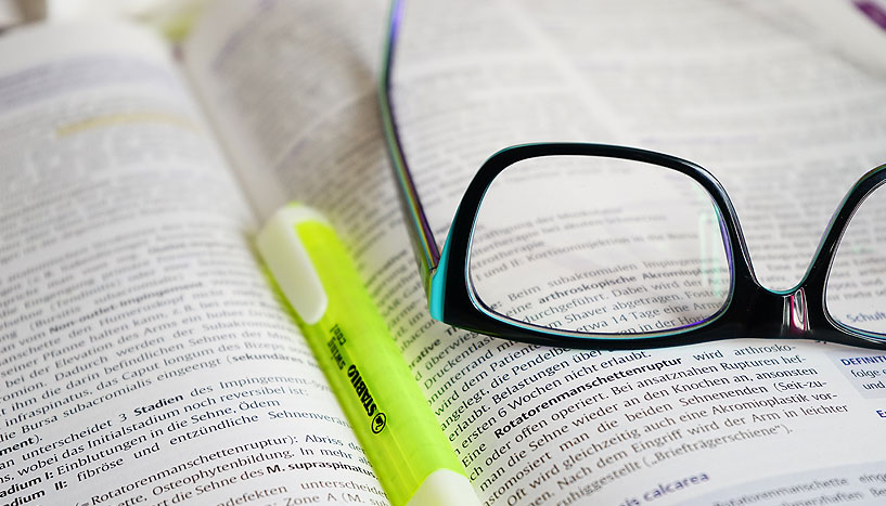 Ein aufgeschlagenes Lehrbuch, auf dem eine Brille und ein neongelber Textmarker liegen.